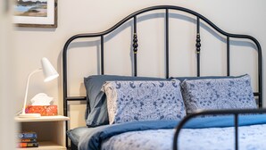 A neatly made bed with a patterned blue bedspread and pillows, a bedside table with a lamp and books, and a framed picture on the wall. - YHZ Cheerful Private Unit 10 min to Airport (ELMSDALE)
