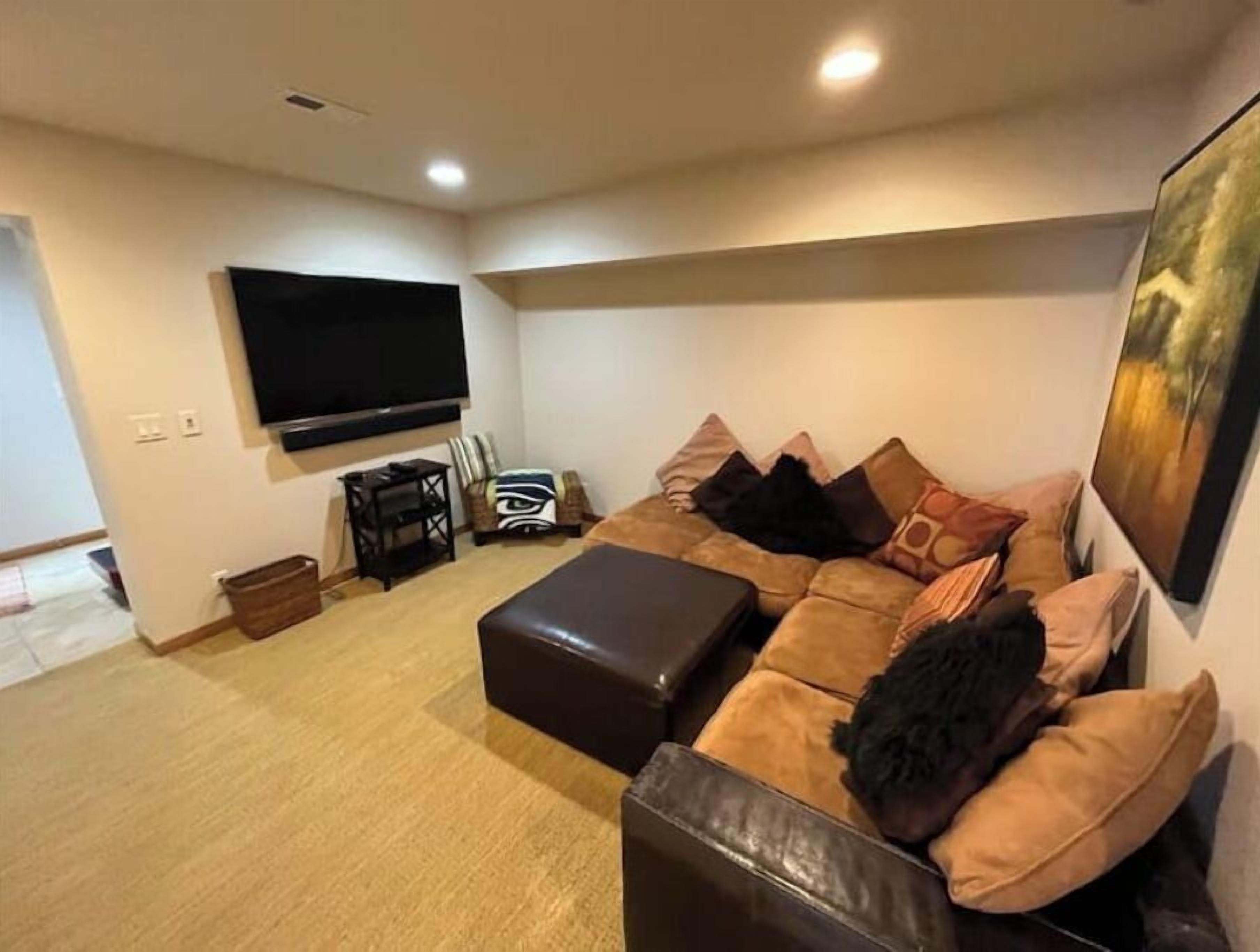 A living room with a large flat-screen TV mounted on the wall, a brown leather sofa with multiple cushions, a small wooden side table, and a basket on the floor.