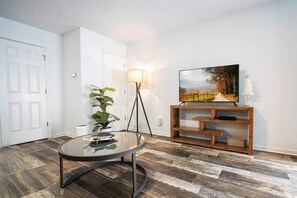 A modern living room with a round glass table, a floor lamp, a TV on a wooden stand, and a potted plant. - Pet-Friendly Smoky Mountain Home w/ Screened Porch (Pigeon Forge)