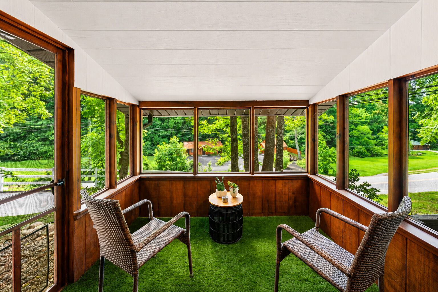A conservatory with two wicker chairs, a small round table, and a view of a green landscape through large windows.