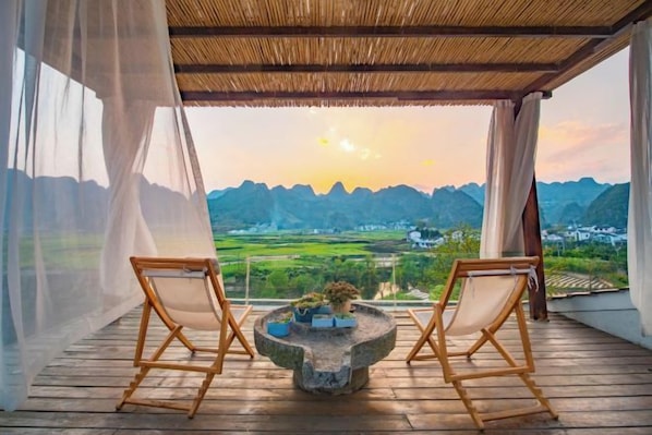 Un balcón con dos sillas de madera, una mesa con una planta y vistas a montañas y campos.
