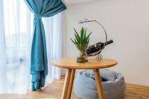 Um quarto com uma mesa redonda de madeira, um vaso de vidro com uma planta, uma garrafa de vinho e um puff azul.