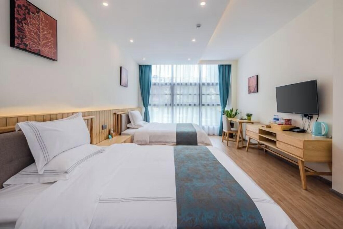 Quarto de hotel com duas camas, uma mesa, uma televisão e uma janela grande com cortinas.