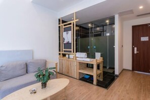 Quarto moderno com sofá, pequena mesa com planta, divisória de madeira e área de chuveiro em vidro.