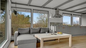 Una cocina moderna con un sofá gris, una mesa de madera y vistas a árboles a través de amplias ventanas.