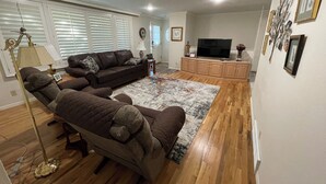 A living room with a sectional sofa, a flat-screen TV, wooden flooring, and a decorative rug.