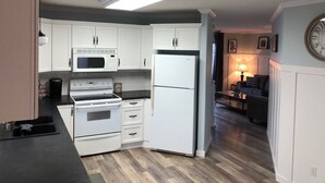 Une cuisine compacte avec des meubles blancs, un four à micro-ondes, une cuisinière et un réfrigérateur. On aperçoit à l’arrière-plan un petit coin repas avec une table et des chaises.