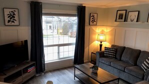 Un salon avec un canapé, une table basse, un téléviseur posé sur un meuble, une lampe et une fenêtre avec des rideaux.
