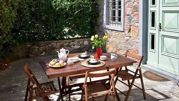 A stone building with a green door, a table set with plates, cups, and a vase of flowers, and a brick archway in the background.