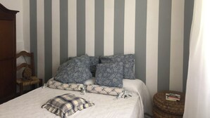 A bedroom with a striped wall, a bed with floral and plaid bedding, a wooden chair, and a small round ottoman with books.
