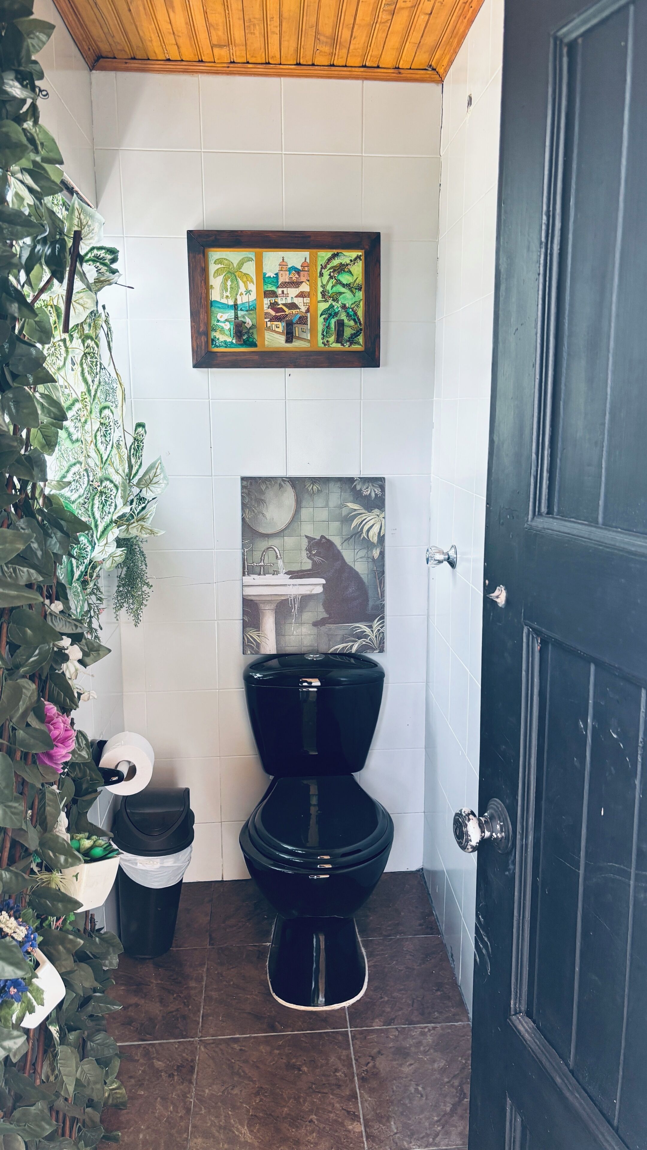 A bathroom with a black toilet, a sink, a framed painting above the sink, and a door to the right.