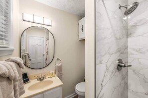 A bathroom with a round mirror, a sink, a toilet, and a shower with a marble wall.
