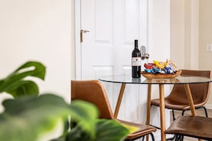 Zona de menjador amb taula de vidre, ampolla de vi, aperitius i una porta al fons.