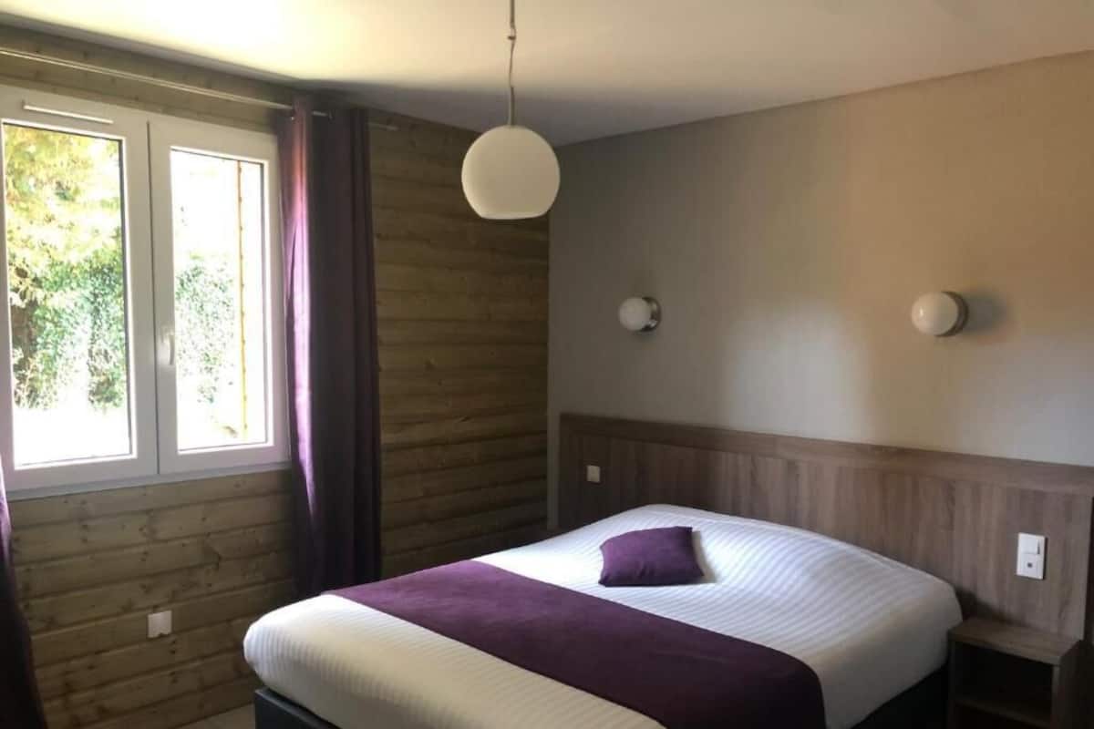 A bedroom with a bed, a window with curtains, a wall-mounted light, and a wooden headboard.