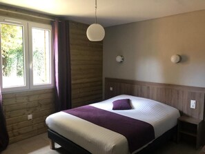 A bedroom with a bed, a bedside table, a hanging light fixture, a window with curtains, and a wooden wall. - Logis Hotel le Val Sarah (Bardouville)