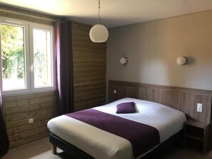 A bedroom with a bed, a window with curtains, a wall-mounted light, and a wooden headboard. - Logis Hotel le Val Sarah (Bardouville)