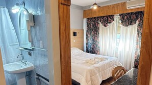 A hotel room with a wooden floor, a bed with white bedding, a bathroom with a sink and mirror, and a window with floral curtains. - Diplomat Hotel  (Buenos Aires)