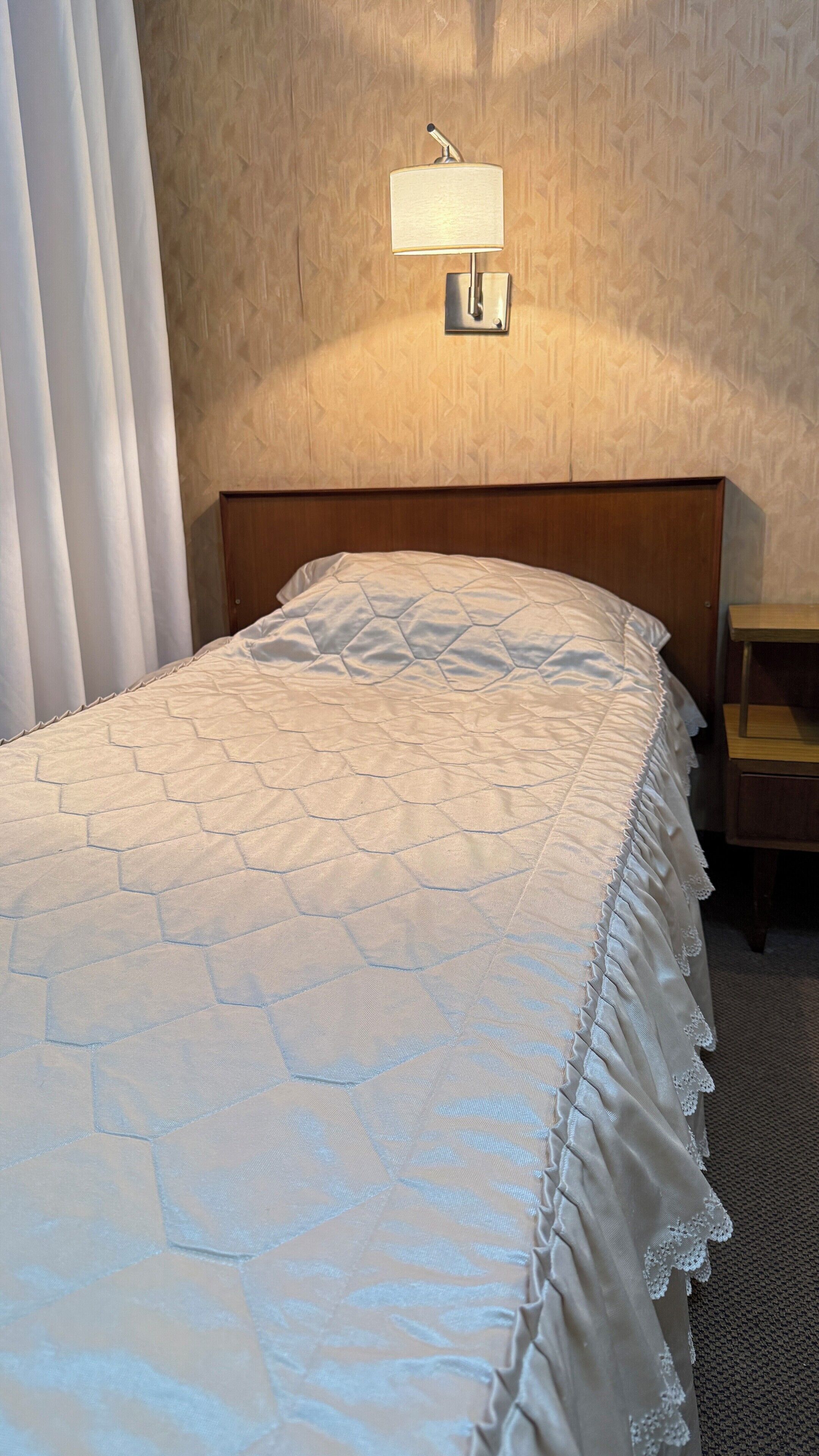 A hotel room with a neatly made bed, a nightstand, and a wall-mounted lamp.