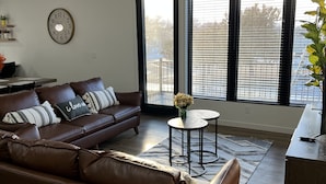 A living room with a brown leather sofa, a round coffee table, a ceiling fan, and a large window with blinds. - LuXury ZeN ZoNE (No Cleaning fee, King size bed). (Omaha)
