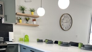 A modern kitchen with a white countertop, grey cabinets, and a green pot in a drawer. There are two pendant lights, a clock, and a shelf with plants. - LuXury ZeN ZoNE (No Cleaning fee, King size bed). (Omaha)