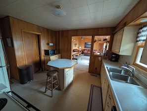 A compact kitchen with wooden walls, a central island, and a dining area visible through an open doorway. - Your lakeside oasis awaits: perfect for families and fun! (Hackensack)