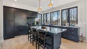 A modern kitchen with dark cabinetry, a large island with bar stools, pendant lighting, and large windows offering a view of trees.