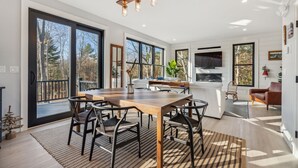 A modern kitchen with a large island, a dining area with a wooden table and chairs, and large windows offering views of the outdoors.