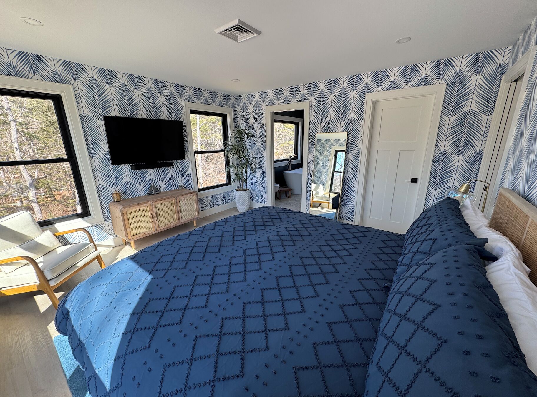 A bedroom with a large bed, two windows, a TV, a plant, and a bathroom visible through an open door.