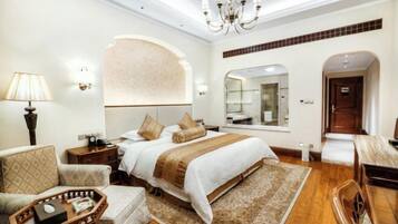 A hotel room with a large bed, a sitting area with a chair and a small table, a chandelier, and a mirror.
