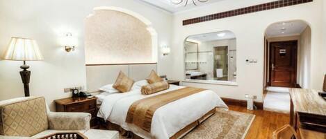 A hotel room with a large bed, a sitting area with a chair and a small table, a chandelier, and a mirror.