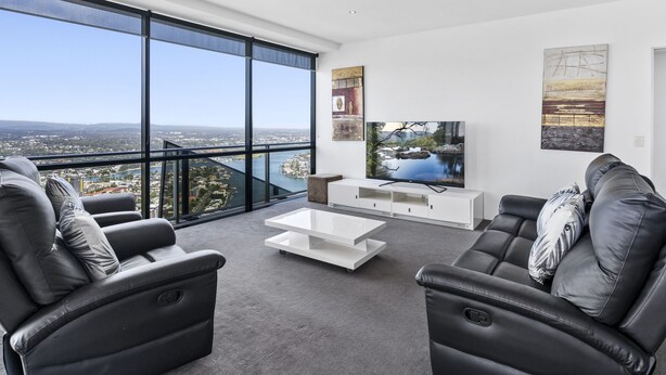A modern living room with a large window offering a city view, a flat-screen TV, and a minimalist white coffee table. - 53rd Floor - Sky High Apartment in Circle on Cavil (Gold Coast)