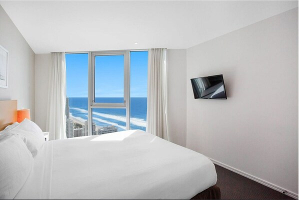 Hotelkamer met een groot bed, een aan de muur bevestigde televisie en uitzicht op de oceaan door middel van kamerhoge ramen.