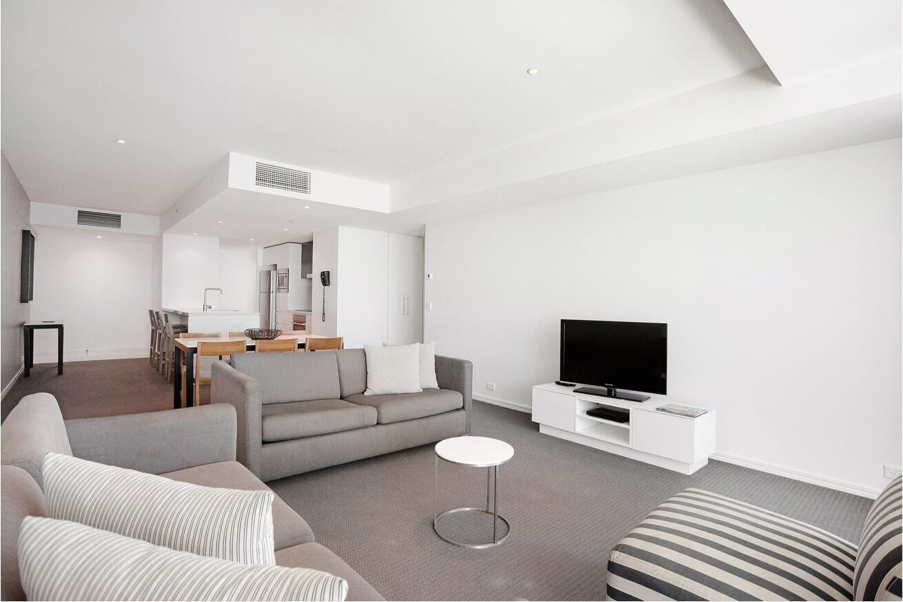 Una sala de estar moderna con un sofá gris, un sillón a rayas, una mesita de centro redonda, un televisor de pantalla plana sobre un soporte blanco, y un comedor al fondo.