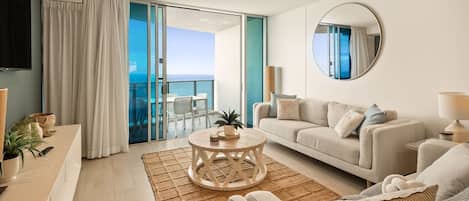 Una sala de estar moderna con un sofá, una mesa de centro y un televisor. Hay un ventanal corredizo que da a un balcón con vistas al mar.