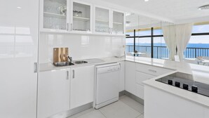A modern kitchen with white cabinets, a dishwasher, and a view of the sea through large windows. - Luxury Oceanview Apartment with Balcony (Surfers Paradise)