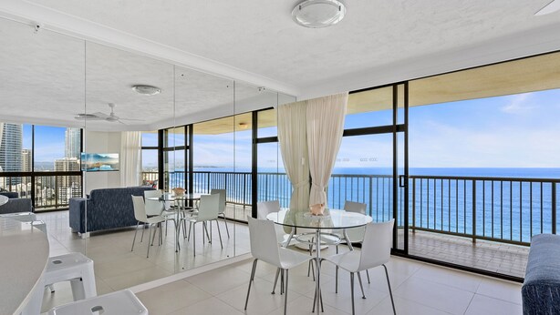 A modern living room with a balcony overlooking the sea, featuring a round dining table, white chairs, and a blue sofa. - Luxury Oceanview Apartment with Balcony (Surfers Paradise)