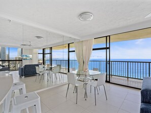 Un salon moderne avec un balcon donnant sur la mer, comprenant une table à manger ronde, des chaises blanches et un canapé bleu.