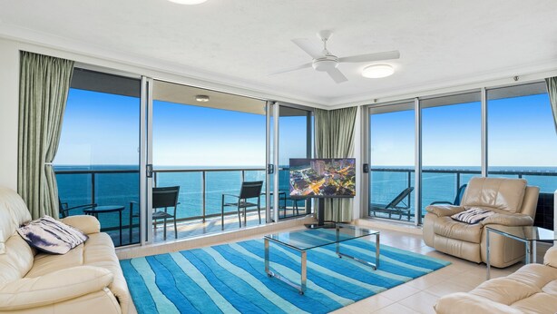 A spacious living room with a large TV, comfortable seating, a glass coffee table, and a view of the ocean.