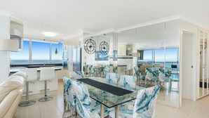 A modern kitchen with a view of the sea, a dining area with a glass table, and a living area with a sofa.