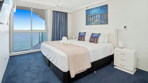 A hotel room with a large bed, a nightstand, a painting, and a view of the sea.