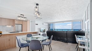 A modern kitchen with a glass dining table and chairs, a black leather sofa, and a view of the sea through large windows. - 28th Floor Oceanfront Bliss in Surfers Paradise (Surfers Paradise)