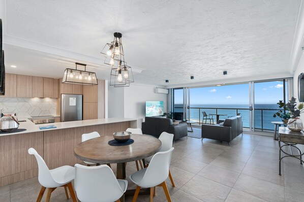 A modern kitchen with wooden cabinets, a central island, and a dining area with white chairs. There is a large window offering a view of the ocean.