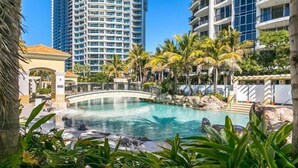 A resort pool area with a bridge, palm trees, and modern buildings in the background. - Sleeps 7 - High Level Ocean View Apt - Chevron! (Surfers Paradise)