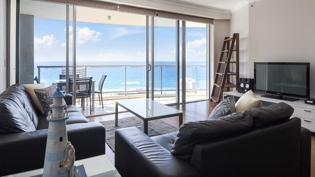 A living room with a view of the ocean, a sofa, a coffee table, a TV, and a bookshelf. - Sleeps 7 - High Level Ocean View Apt - Chevron! (Surfers Paradise)