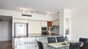 A modern kitchen with a central island, a dining area with a glass table and black chairs, and a kitchen island with a sign. - Sleeps 7 - High Level Ocean View Apt - Chevron! (Surfers Paradise)