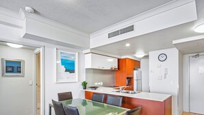 Een moderne keuken met witte kasten, een oranje accentmuur, een roestvrijstalen koelkast, en een eethoek met een glazen tafel en lederen stoelen.