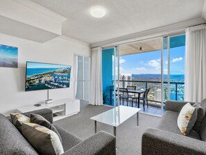 A modern living room with a large TV, comfortable seating, and a balcony with a city view. - Level 29 Apt - Towers of Chevron - 2 Bed 2 Bath (Surfers Paradise)