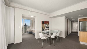 Une chambre d’hôtel moderne avec balcon, un coin repas doté d’une table en verre et de chaises blanches, et une kitchenette.