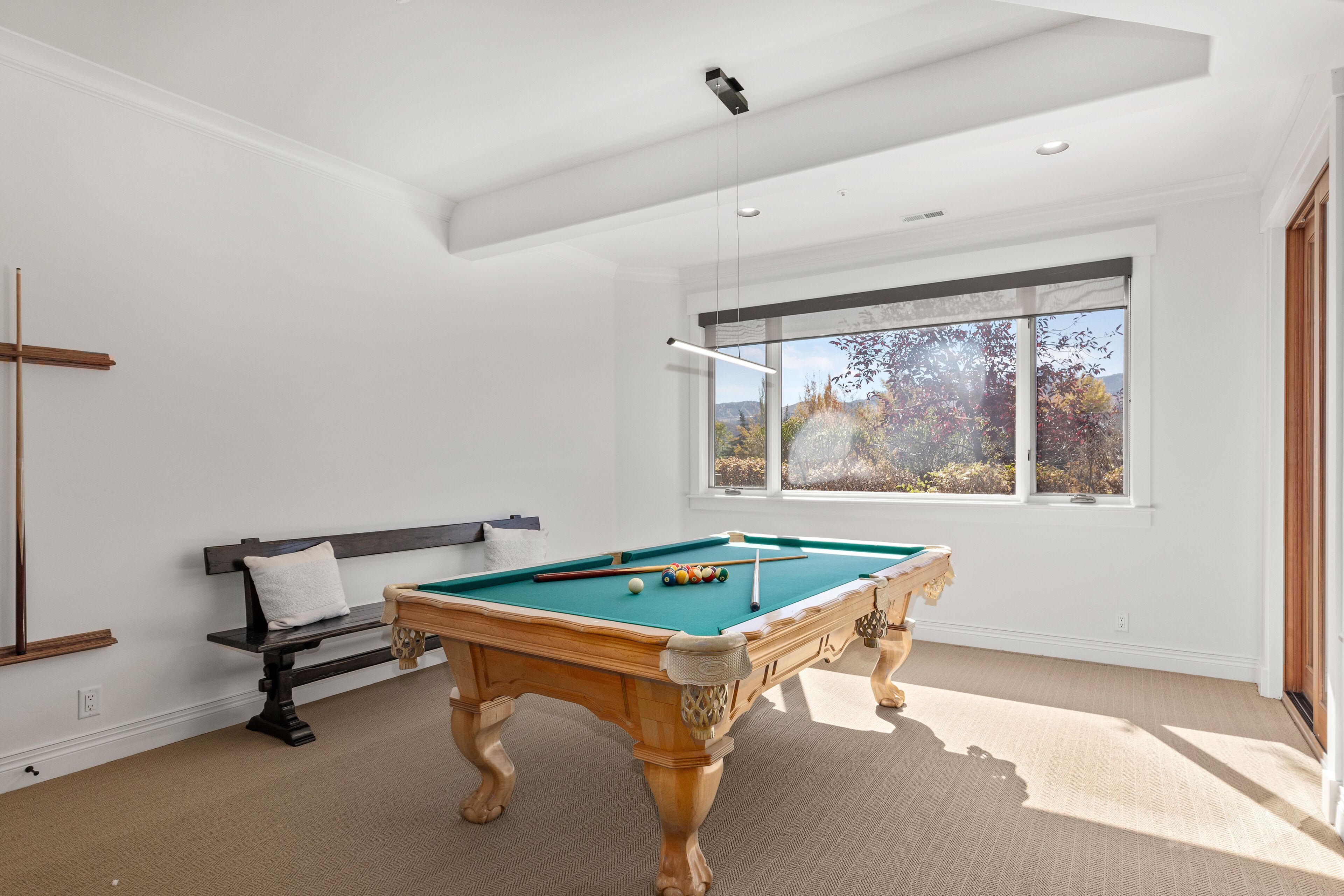 Una mesa de billar en una habitación con un ventanal grande, un banco y una viga de madera en la pared.