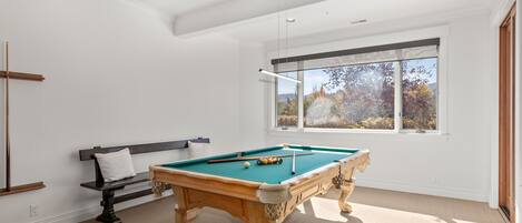Una mesa de billar en una habitación con un ventanal grande, un banco y una viga de madera en la pared.